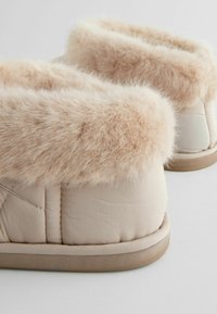 Beige tøfler med imitation pelsfór, quiltet tekstur, blød og luksuriøs krave samt en holdbar gummisål.