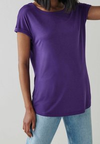 Lila Kurzarm-T-Shirt mit Rundhalsausschnitt und aufgeschlagenen Ärmeln, aus einem glatten Stoff gefertigt. Kombiniert mit hellblauen Jeans.