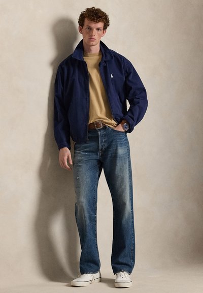 Navy blue jacket, beige shirt, distressed blue jeans, and white sneakers. Model stands with arms crossed, showcasing casual ensemble.
