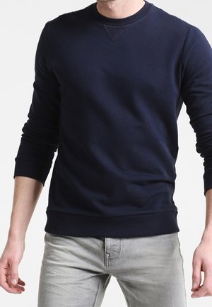 Uomo che indossa una felpa a maniche lunghe con scollo a girocollo blu navy e jeans grigio chiaro, in piedi davanti a uno sfondo chiaro e uniforme.