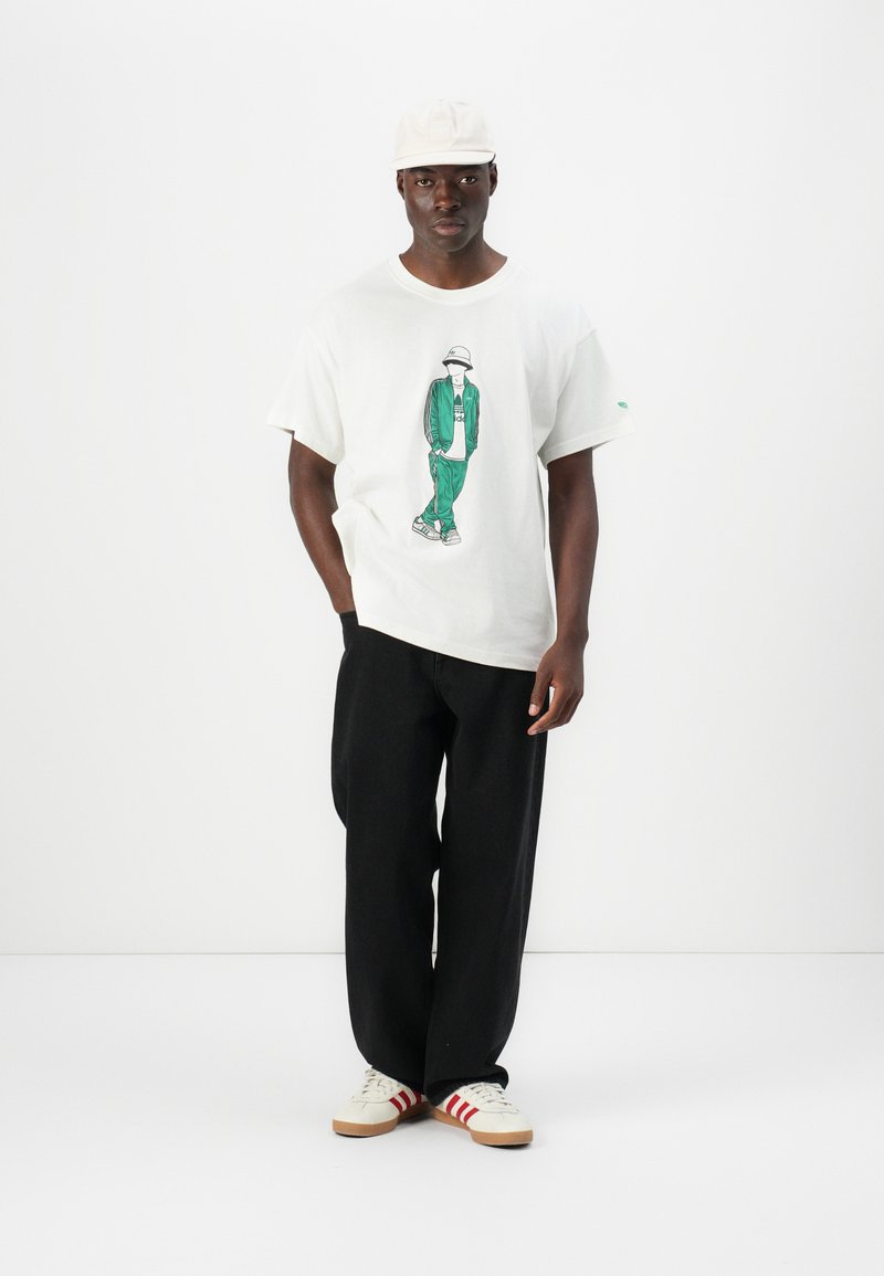 Biała bawełniana koszulka z zielonym nadrukiem, w połączeniu z czarnymi luźnymi spodniami i białymi trampkami z czerwonymi paskami.