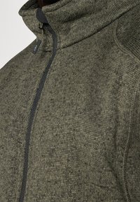 Khakifarbene Fleecejacke mit Reißverschluss, hohem Kragen, strukturiertem Stoff, kontrastierendem schwarzen Reißverschluss und dezenten Sprenkeln.