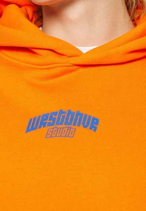 Personne portant un sweat à capuche orange vif, un pantalon marron foncé retroussé et des baskets vertes et blanches, debout à l'intérieur près d'une fenêtre avec un mur en béton.
