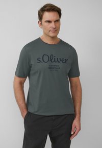 Szara bawełniana koszulka z okrągłym dekoltem i krótkimi rękawami. Na przodzie znajduje się granatowy nadruk "s.Oliver Elevated Essentials".