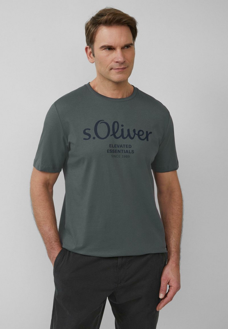 Szara bawełniana koszulka z okrągłym dekoltem i krótkimi rękawami. Na przodzie znajduje się granatowy nadruk "s.Oliver Elevated Essentials".