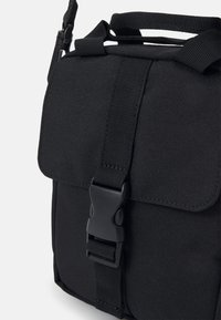 Black fabric crossbody bag with a flap closure, adjustable strap, and a plastic buckle. Features a textured surface and robust stitching.