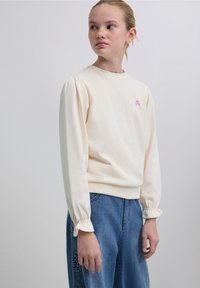 Hanorac crem cu mâneci lungi bufante și tiv cu dungi, având un mic desen roz pe piept, asortat cu blugi de culoare albastru deschis.