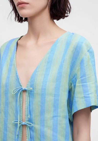 Femme portant un haut rayé bleu et vert à manches courtes avec deux fermetures à nouer à l'avant et un profond décolleté en V.