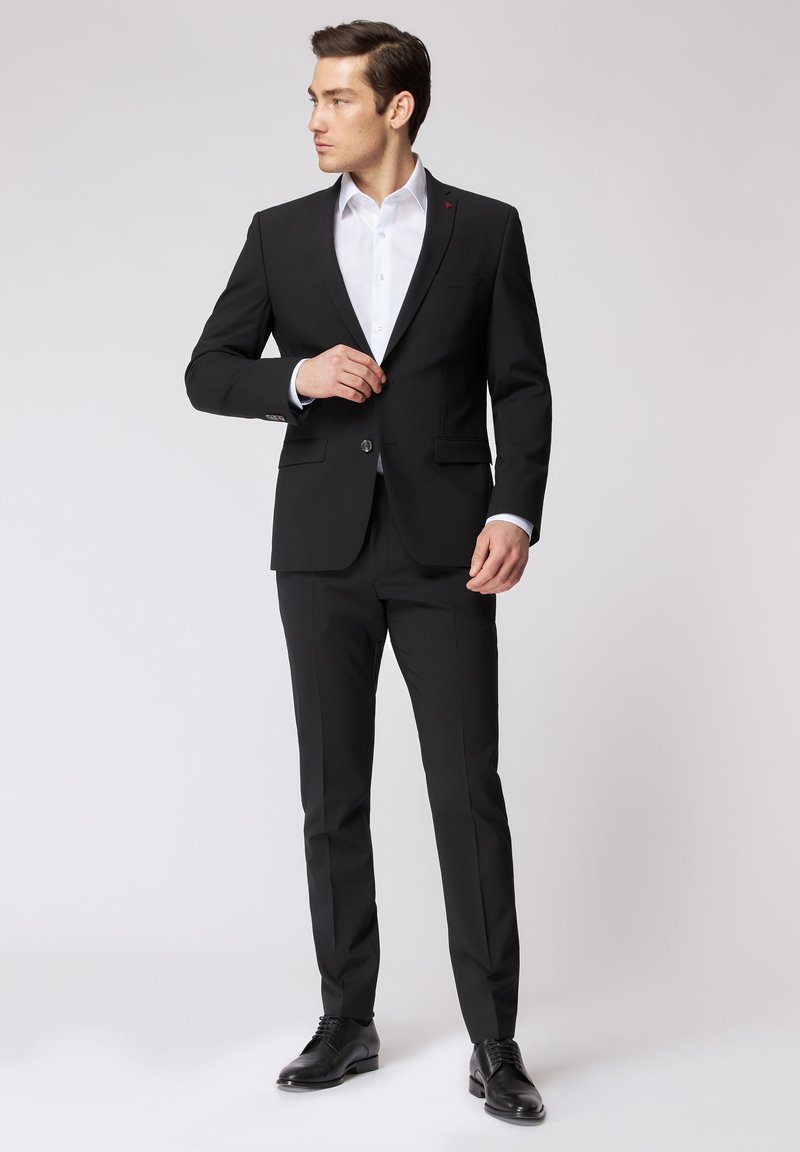 Completo nero con una vestibilità su misura, design monopetto e revers a scialle. Camicia bianca sotto, abbinata a scarpe in pelle nere.