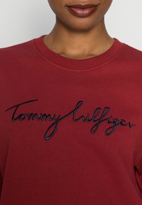 Sudadera roja de cuello redondo, con el logotipo bordado de "Tommy Hilfiger" en azul marino. Textura suave y cuello acanalado.