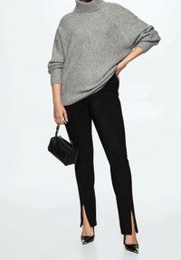 Grau gestrickter Rollkragenpullover mit übergroßer Passform, kombiniert mit schwarzen, maßgeschneiderten Hosen mit seitlichen Schlitzen und spitzen schwarzen High Heels. Schwarze Handtasche inklusive.