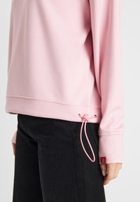 Pink sweatshirt with long sleeves, drawstring at the hem, ribbed cuffs, and a small red tag on the sleeve. Smooth fabric texture.
