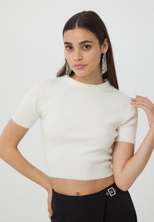 Maglione corto bianco a coste con maniche corte e scollo tondo, abbinato a una gonna nera con dettaglio di fibbia metallica.