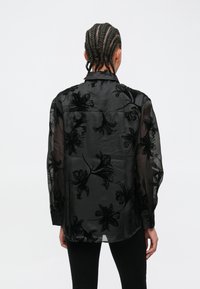 Čierna priehľadná blúzka s kvetinovou výšivkou, dlhými rukávmi a golierom. Má voľný strih a gombíkové detaily na manžetách.