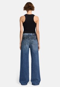 Weite blaue Jeans im used-Look mit Metallnieten, kombiniert mit einem schwarzen Tanktop. Der Stoff ist Denim mit einer strukturierten Oberfläche.