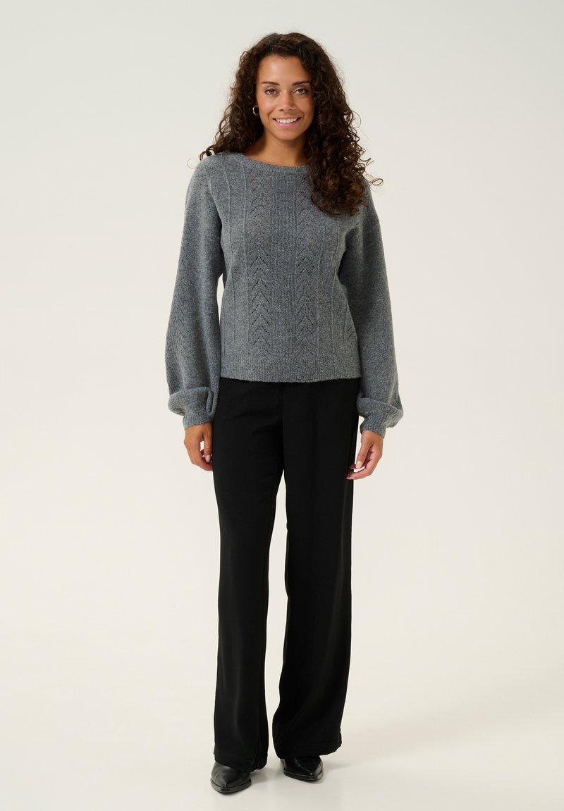 Grauer Strickpullover mit strukturiertem Muster, rundem Halsauschnitt und puffärmeligen Ärmel, kombiniert mit schwarzen weit geschnittenen Hosen und schwarzen Schuhen.