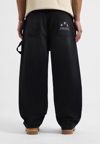 Pantalones de mezclilla negros de corte holgado con un ajuste relajado. Presentan dos bolsillos traseros con un diseño de estrella y texto. Acentuados con una correa lateral.