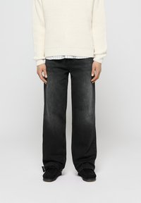 Pantalones vaqueros anchos negros de denim, con un acabado desgastado. Combinados con un suéter texturizado en color crema, sobre una camisa a rayas.