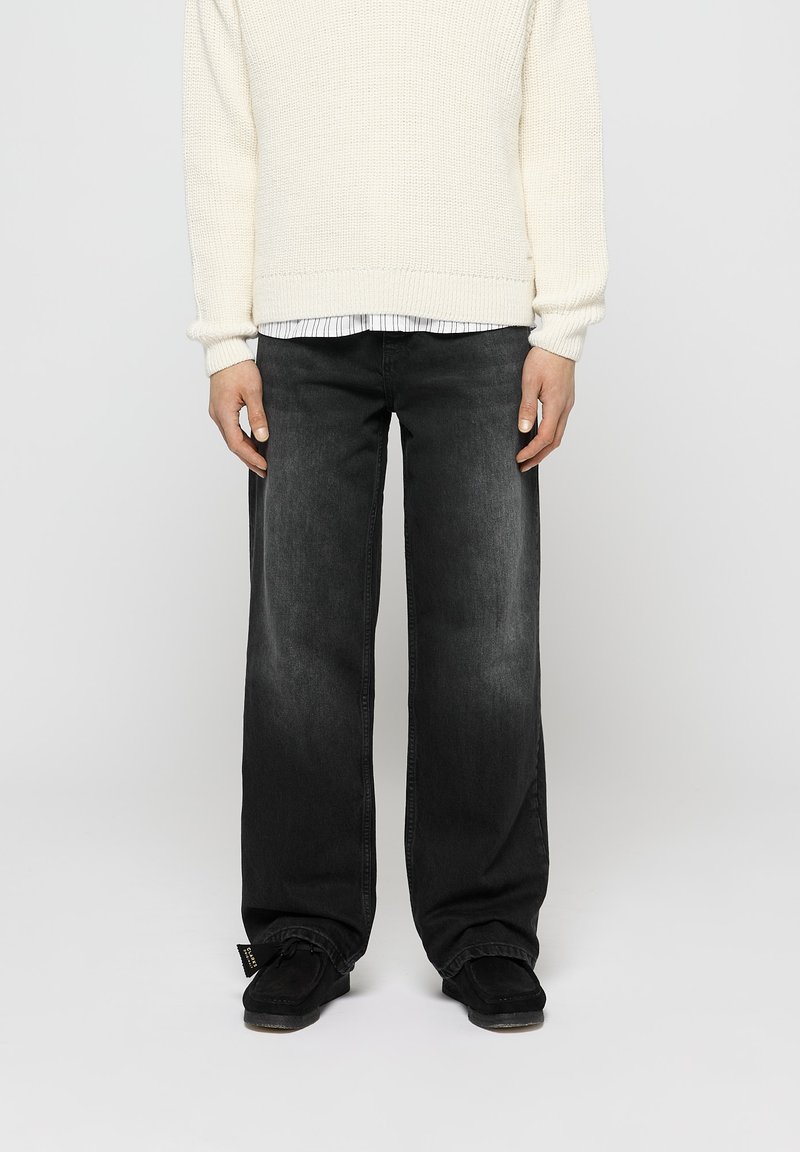 Pantalones vaqueros anchos negros de denim, con un acabado desgastado. Combinados con un suéter texturizado en color crema, sobre una camisa a rayas.