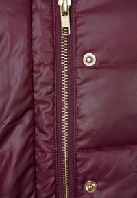 Primo piano di una giacca imbottita color borgogna con cerniera dorata e bottoni a pressione, con tessuto liscio e leggermente lucido.