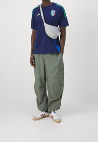 Granatowa koszulka Adidas z zielonymi i niebieskimi akcentami, szara torba na ramię, jasnoszare spodnie cargo oraz białe sneakersy z czerwonymi paskami.