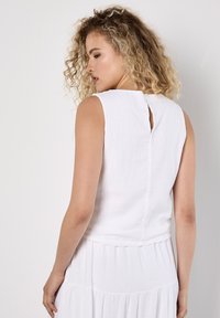 Een vrouw met krullend haar kijkt iets weg, gekleed in een mouwloos wit topje met een keyhole rug, en een wijdvallende witte rok.