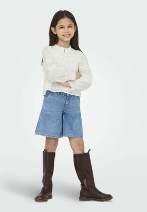 Blouse blanche à manches longues avec des accents texturés, short en denim bleu, et bottes en cuir marron jusqu'aux genoux. Bras croisés, souriante.