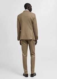 Completo marrone con texture, caratterizzato da una giacca a singolo spacco e pantaloni su misura, abbinato a una camicia bianca e scarpe nere.