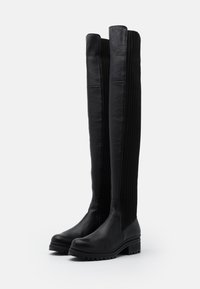 Polven korkeuteen yltävät mustat nahkasaappaat, joissa on ribbineulosta, pyöreät kärjet ja massiiviset kumipohjat. Saappaissa on sileä pinta ja pystysuorat tekstuurikoristeet.