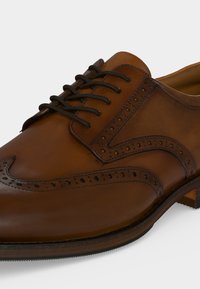 Chaussure habillée pour homme en cuir marron avec détails brogue à bout golf et lacets noirs, vue en gros plan sous un angle.