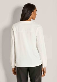 Vrouw met lang donker haar, gekleed in een eenvoudige witte blouse met lange mouwen en zwarte broek, staande met haar rug naar de camera tegen een neutrale achtergrond.