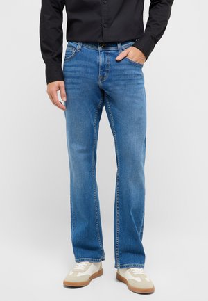 Mann in blauer Jeans, schwarzem Hemd mit Knöpfen und weißen Turnschuhen, der mit einer Hand in der Tasche vor einem schlichten Hintergrund steht.