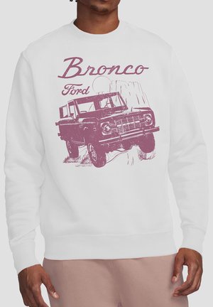 Mann in weißem Sweatshirt mit Vintage-Grafik eines violetten Ford Bronco-Trucks, der felsiges Gelände erklimmt.