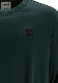 Mörkgrön långärmad tröja med rund halsringning, prydd med en fyrkantig logotyp med en orange design på vänster bröst. Mjuk tygstruktur.