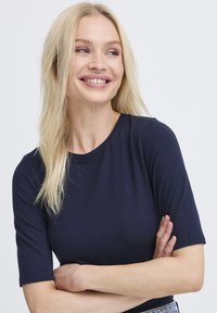 Marineblaue, taillierte T-Shirt aus weichem Stoff mit kurzen Ärmeln und rundem Ausschnitt, getragen mit verschränkten Armen und neutralem Hintergrund.