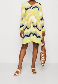 Vestido de manga corta y escote en V en un patrón de tie-dye verde, amarillo y naranja, con una silueta acampanada y una tela ligera. Lleva sandalias beiges y un bolso de mimbre.