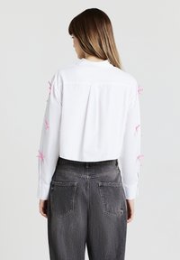 Weißes kurz geschnittenes Hemd mit langen Ärmeln, verziert mit rosa Bandakzenten an den Ärmeln und einer Rückenfalte, kombiniert mit hoch taillierter grauer Hose.