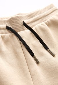 Pantalones de chándal beige con cintura acanalada, que cuentan con cordones negros y puntas de metal marcadas "BAKER". Textura suave, diseño casual.