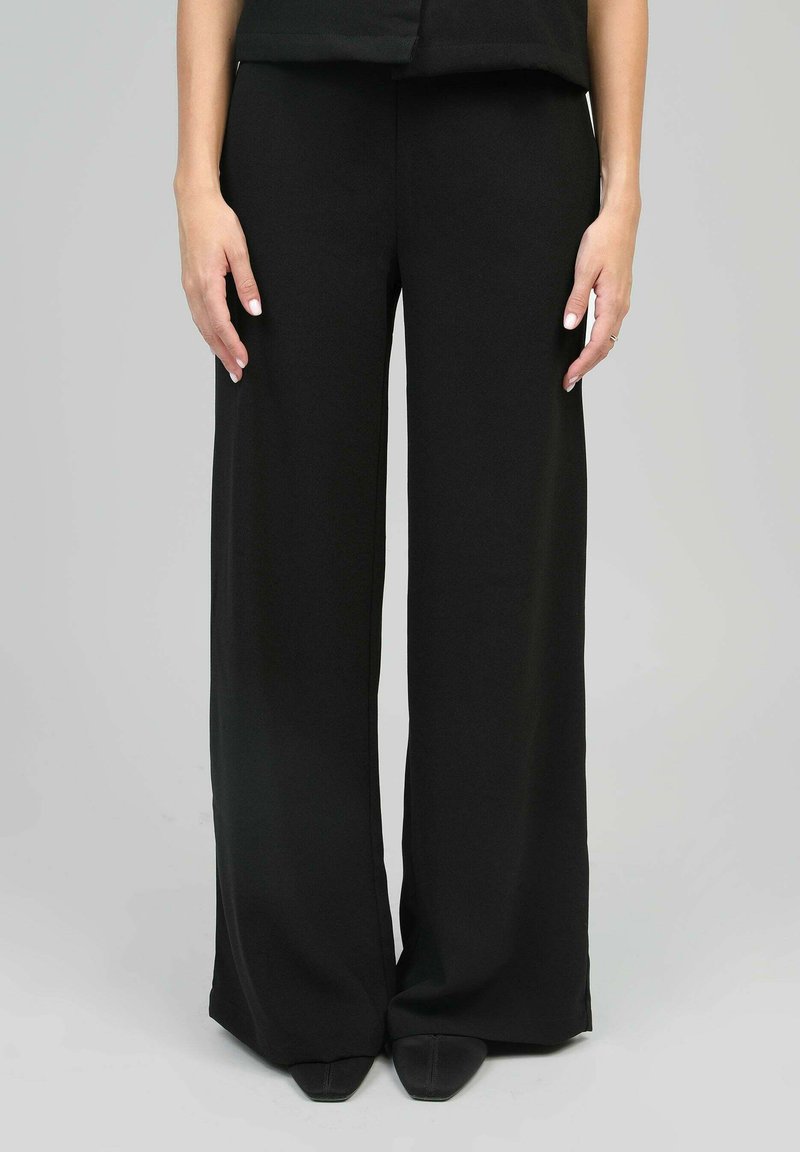 Pantalon noir à jambe large en tissu lisse, doté d'une taille haute, de lignes épurées et de plis subtils pour une structure ajoutée.