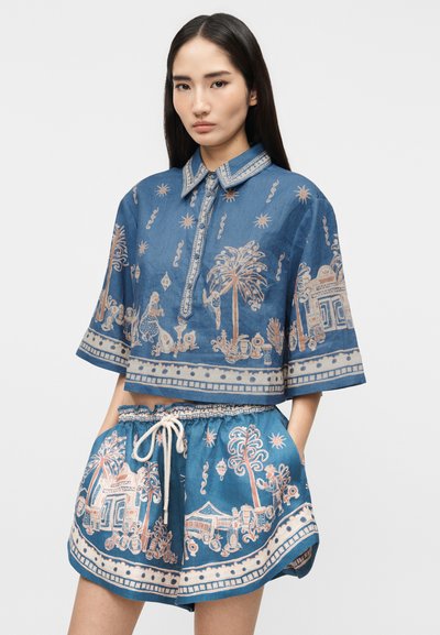 Γυναίκα που φοράει μπλε κοντό πουκάμισο με γιακά και ταιριαστά σορτς με μπεζ περίπλοκα σχέδια από φοίνικες, ανθρώπους και κτίρια.