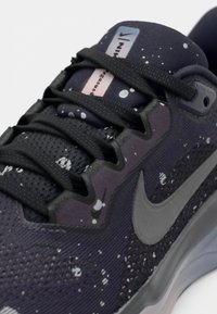 Gros plan sur une chaussure de course Nike noire avec un motif moucheté blanc, des lacets noirs et un logo Nike gris sur le côté.