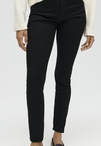 Strakke zwarte broek gedragen door een persoon met een lichte huid, gecombineerd met een wit top met lange mouwen en zwarte instapschoenen.