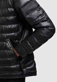 Jachetă neagră puffer cu o textură lucioasă, având quilturi orizontale, un buzunar cu fermoar și un logo de brand pe umăr.