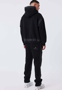 Hombre vestido con una sudadera con capucha negra y pantalones de chándal negros con un logo en el bolsillo trasero, de pie y mirando hacia un lado, con zapatillas blancas.