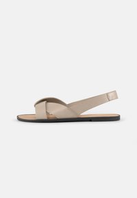 Beige, flate sandal laget av mykt materiale, med kryssede stropper og slingback-design. Svart såle med minimal mønstring.