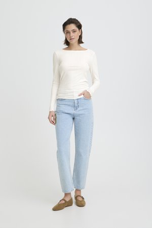 Vrouw staand met hand in zak, gekleed in een witte top met lange mouwen, lichtblauwe jeans en bruine platte schoenen tegen een effen achtergrond.