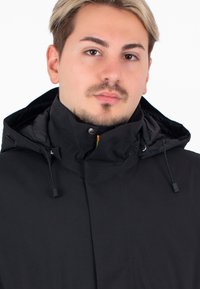 Giacca nera con cappuccio e colletto alto, realizzata in un tessuto liscio. Presenta cordini e chiusura a pressione sul collo.