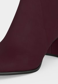 Gros plan d’une botte d’ cheville bordeaux avec un talon bloc et une texture cuir lisse sur fond blanc.