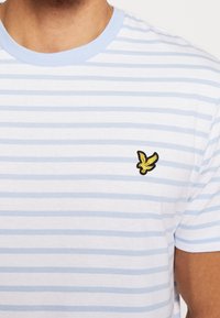 Camiseta de rayas horizontales azul claro y blanca con un cuello azul claro y un pequeño logo de un pájaro amarillo en el lado izquierdo del pecho.