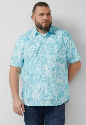 Mężczyzna z brodą, ubrany w jasnoniebieską, kwiatową, krótki rękaw, zapinaną na guziki koszulę i ciemne dżinsy, stoi na tle gładkiego białego tła.